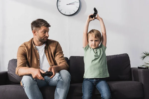 QUIIV, UCRÂNIA - JUNHO 9, 2020: menino bonito segurando joystick e mostrando gesto vencedor perto pai chateado — Fotografia de Stock