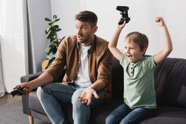 KYIV, UCRANIA - 9 de junio de 2020: niño emocionado sosteniendo el joystick y mostrando un gesto ganador cerca de un padre confuso mostrando un gesto de encogimiento - foto de stock