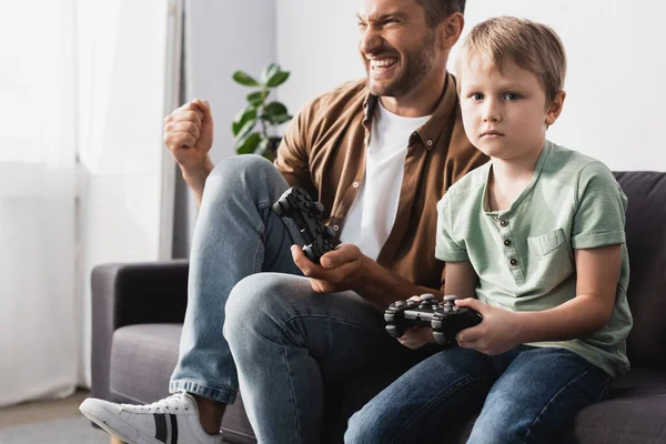 KYIV, UKRAINE - JUNE 9, 2020: upset boy holding joystick and looking at camera near excited father showing winner gesture — Stock Photo