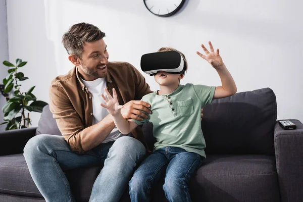 Souriant père touchant fils surpris en utilisant vr casque et gestuelle tandis que assis sur le canapé — Photo de stock