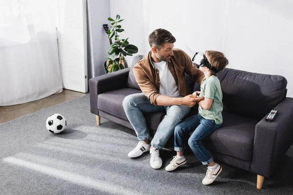 Высокий угол зрения мужчины, трогающего сына с помощью гарнитуры VR, сидя на диване — стоковое фото