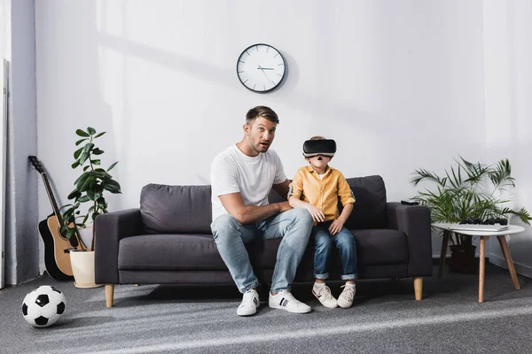 Bel homme et fils excité dans casque assis sur le canapé à la maison — Photo de stock