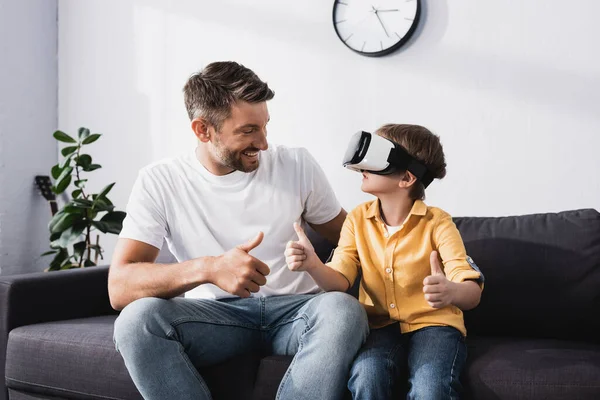 Smiling man and son in vr headset showing thumbs up while sitting on sofa — Stock Photo