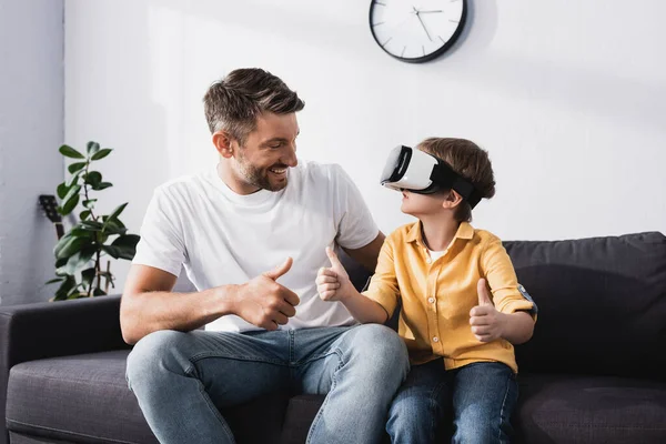 Sorrindo homem e filho em vr fone de ouvido mostrando polegares para cima enquanto sentado no sofá — Fotografia de Stock