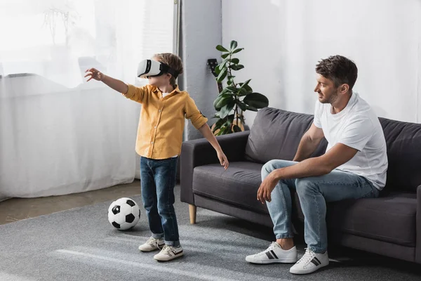 Boy in vr headset standing with outstretched hand near father sitting on sofa — Stock Photo
