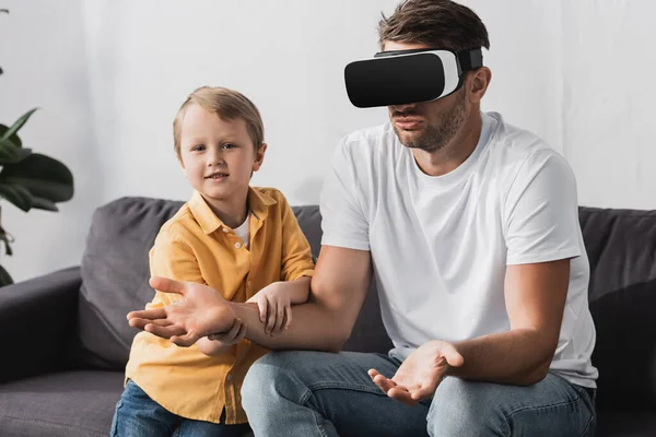 Lindo chico tocando mano de padre en vr auriculares mostrando gesto encogiéndose de hombros - foto de stock