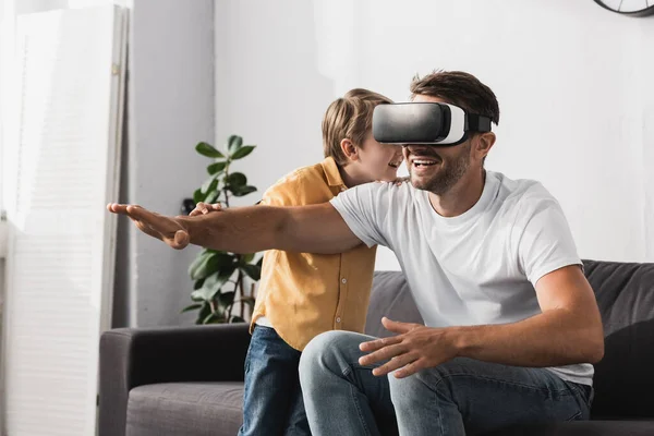 Lächelnder Mann im vr-Headset sitzt mit ausgestreckter Hand in der Nähe seines entzückenden Sohnes — Stockfoto