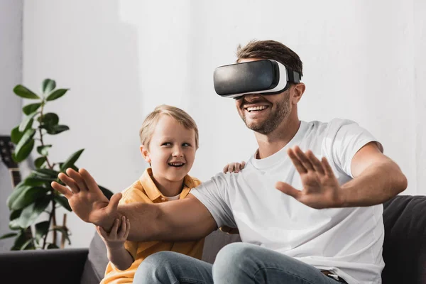 Sonriente padre en vr auriculares gesto mientras lindo hijo sentado en sofá - foto de stock