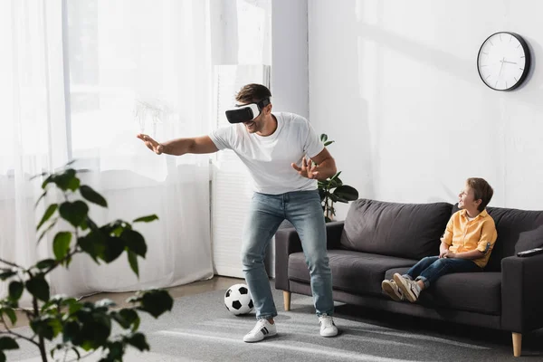 Selective focus of man in vr headset walking and gesturing while adorable son sitting on sofa — Stock Photo