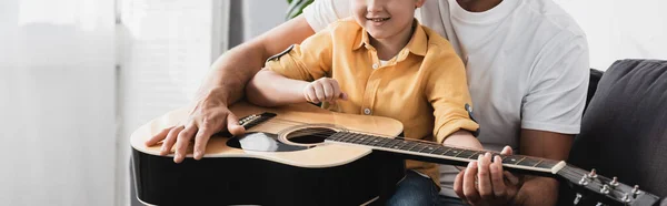 Visão cortada do menino sentado de joelhos pais e aprendendo a tocar guitarra acústica — Fotografia de Stock