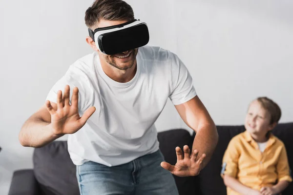 Messa a fuoco selettiva di uomo sorridente in auricolare vr gesticolare mentre adorabile figlio seduto sul divano — Foto stock