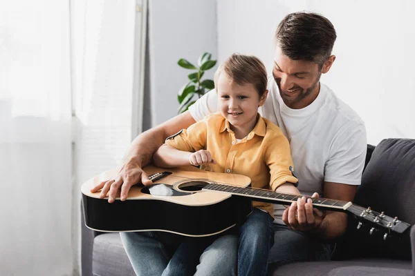 Lächelnder Junge, der auf den Knien des Vaters sitzt und Akustikgitarre spielen lernt — Stockfoto