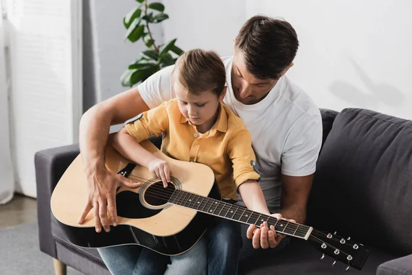 Cute boy sitting of fathers knees and learning how to play acoustic guitar — Stock Photo