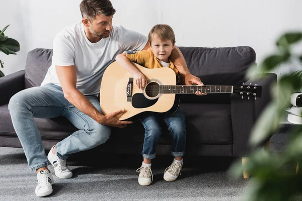 Enfoque selectivo de padre guapo enseñando hijo cómo tocar la guitarra acústica - foto de stock