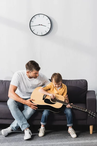 Уважний хлопчик вчиться грати на акустичній гітарі, сидячи на дивані біля батька — стокове фото