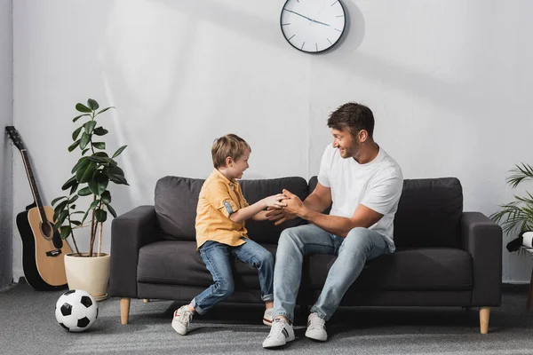 Alegre padre e hijo chistosamente luchando en el sofá cerca de macetas de plantas, pelota de fútbol y guitarra - foto de stock
