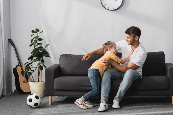 Отец и очаровательный сын в шутку дерутся на диване рядом с футбольным мячом и растением в горшках — стоковое фото