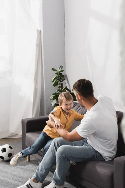 Padre y feliz hijo chistosamente luchando en el sofá cerca de la pelota de fútbol - foto de stock