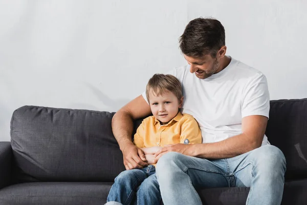 Glücklicher Vater umarmt entzückenden Sohn, während er zu Hause auf dem Sofa sitzt — Stockfoto