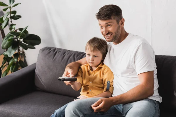 Fröhlicher Vater und gelangweilter Sohn, der während des Fernsehens Kanäle mit Fernbedienung anklickt — Stockfoto