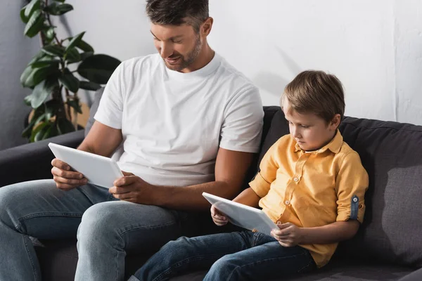 Père souriant et fils concentré assis sur le canapé et en utilisant des tablettes numériques — Photo de stock