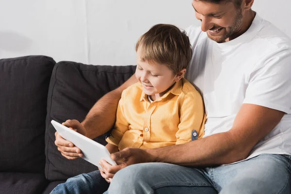 Feliz padre e hijo sonriente sentado en el sofá y el uso de la computadora portátil juntos - foto de stock