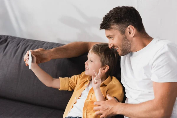 Glücklicher Junge zeigt Siegesgeste, während er mit lächelndem Vater das Smartphone in die Hand nimmt — Stockfoto