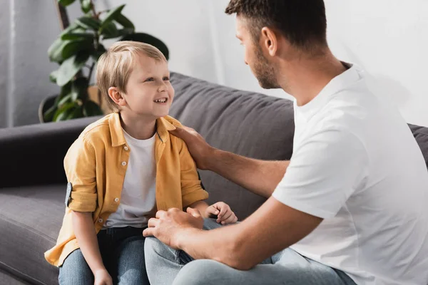 Батько торкається плеча усміхненого сина, розмовляючи з ним на дивані — стокове фото