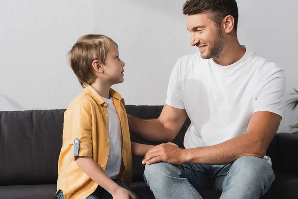 Smiling father touching cute son while sitting on sofa and talking to him — Stock Photo