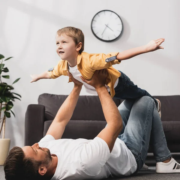 Padre acostado en el suelo y sosteniendo adorable hijo imitando volar con las manos extendidas - foto de stock