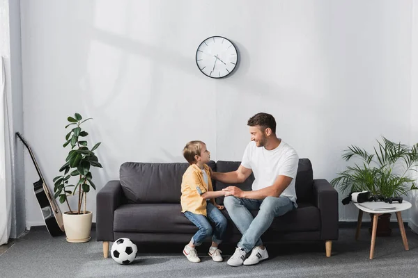 Отец и сын разговаривают, сидя на диване возле футбольного мяча и горшков растений — стоковое фото