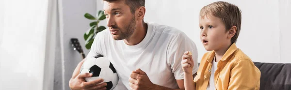 Tiro panorâmico de homem preocupado assistindo tv enquanto segurando bola de futebol perto do filho atencioso — Fotografia de Stock