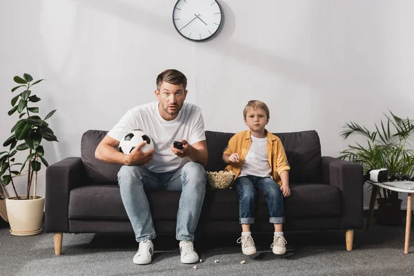 Worried man holding soccer ball and tv remote controllers near son eating popcorn — Stock Photo