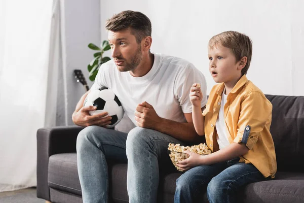 Inquiet père et fils regardant la télévision tout en tenant ballon de football et bol de maïs soufflé — Photo de stock