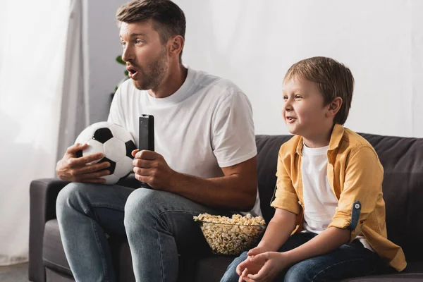 Homem chocado segurando bola de futebol e tv controle remoto perto de filho sorridente — Fotografia de Stock