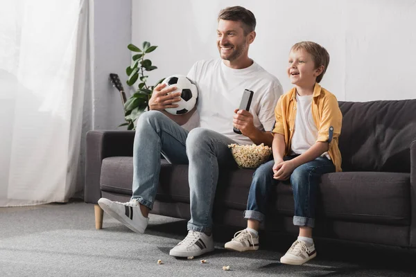 Feliz hombre sosteniendo pelota de fútbol y tv mando a distancia cerca sonriente hijo - foto de stock