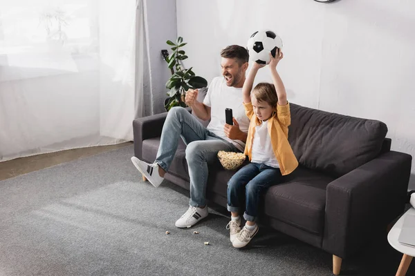 Excited man showing winner gesture near son holding soccer ball in raised hands while watching tv — Stock Photo