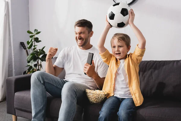 Hombre emocionado mostrando gesto ganador cerca de hijo sosteniendo pelota de fútbol en manos levantadas mientras mira la televisión - foto de stock