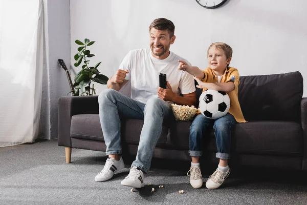 Happy boy holding soccer ball and pointing with finger near excited father while watching tv — Stock Photo