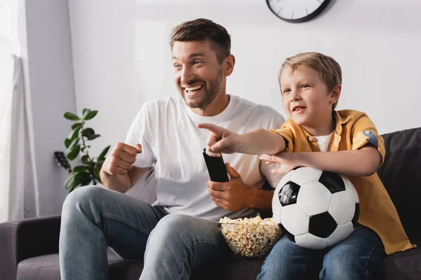 Excited boy holding soccer ball and pointing with finger near cheerful father while watching tv — Stock Photo