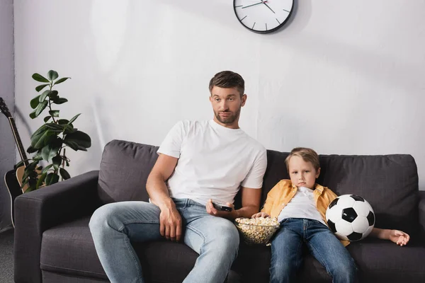 Entediado pai e filho assistindo tv enquanto sentado no sofá com pipocas e bola de futebol — Fotografia de Stock