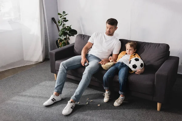 Aburrido padre e hijo sentado en el sofá con palomitas de maíz y pelota de fútbol y viendo la televisión - foto de stock