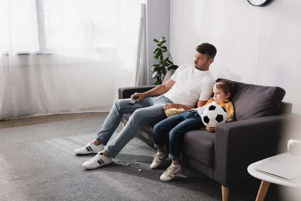 Padre e figlio annoiato a guardare la tv mentre si siede sul divano con popcorn e pallone da calcio — Foto stock