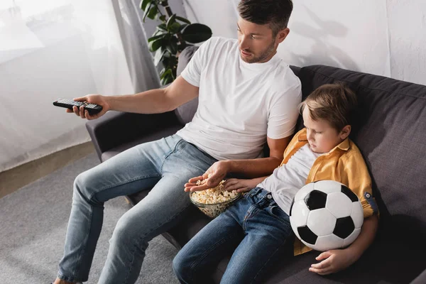 Vista de ángulo alto de niño sosteniendo pelota de fútbol mientras ve la televisión y comer palomitas de maíz con el padre - foto de stock