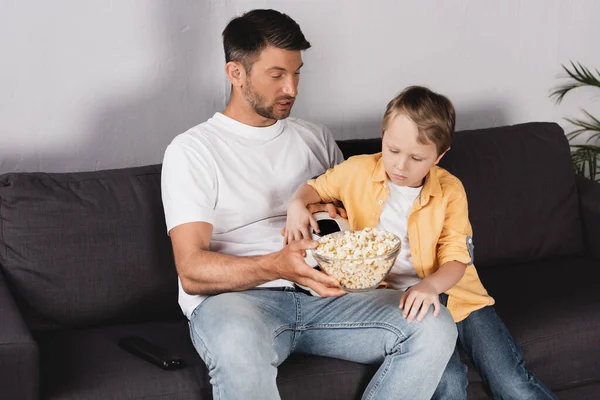 Pai e filho comendo pipocas enquanto sentado no sofá e assistindo tv — Fotografia de Stock