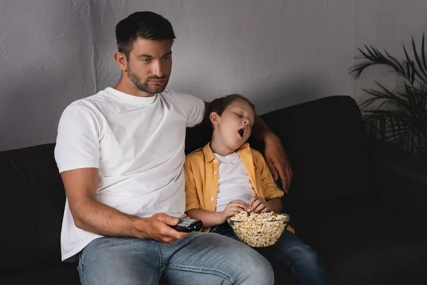 Aburrido padre sosteniendo TV mando a distancia cerca bostezando hijo sosteniendo tazón de palomitas de maíz - foto de stock