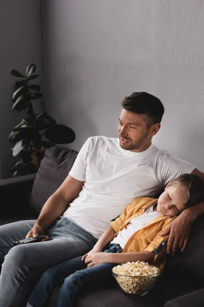 Sorrindo pai assistindo tv perto filho dormindo perto de tigela de pipoca — Fotografia de Stock