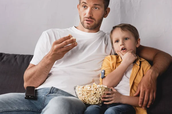 Padre e hijo concentrados viendo la televisión y comiendo palomitas de maíz en el sofá en casa - foto de stock
