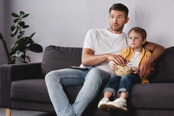 Внимательный отец и сын смотрят телевизор и едят попкорн на диване дома — стоковое фото