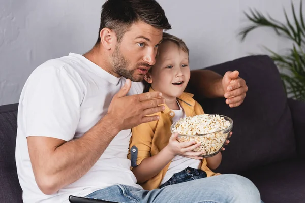 Père concentré et fils souriant regardant la télévision tout en tenant bol de maïs soufflé — Photo de stock